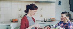 Cheerful happy mother cutting tomatoes while cooking and talking to her smiling daughter feeding mom in the kitchen at home. Family, cook, and people concept